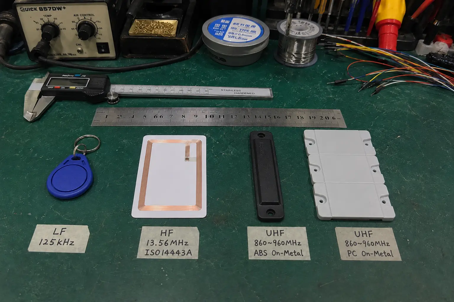Bench comparison of LF 125kHz keyfob, HF 13.56MHz coil card, and UHF ABS and PC on-metal tags with calipers and steel ruler.